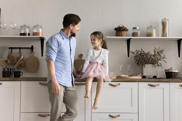 Young dad talking to little daughter in cozy domestic kitchen, spend time together, share pleasant news, recollect, having friendly conversation, smile, enjoy friendship and warm relationships at home