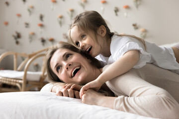 Playful little 4s girl spend playtime on bed with laughing mum, lying together on comfortable mattress, enjoy fresh beddings and good morning, start new day, close up. Family time, carefree childhood