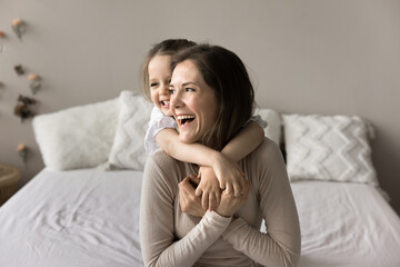 In cozy domestic bedroom laughing awakened little 3s daughter cuddling from behind, piggyback her cheery young mother, enjoy morning together at home. Family ties, happy motherhood, carefree childhood