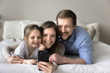 Young couple with little daughter using modern smart phone, look at cellphone screen having pleasant talk via video call application, taking selfie pictures, spend carefree leisure on-line on internet