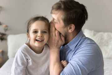 Fototapeta premium Close up shot of young dad whispering secrets to cute little daughter, sit together on bed, share confidential information, express trust and deep connection, having fun, playing in morning at home