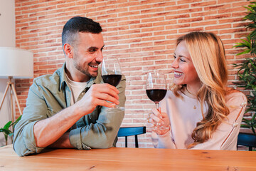 Young caucasian couple smiling and toasting red wine glasses together at home living room. Real affectionate people relaxing drinking a wineglass on a date. Husband and wife bonding indoors.
