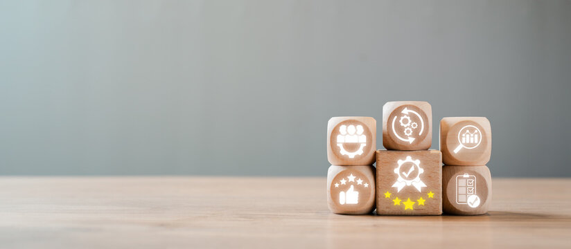 Wooden blocks with icons symbolizing quality management, business excellence, and continuous improvement on a wooden table with a grey background.