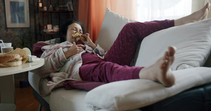 Unkept Lazy woman lying on the sofa, eating fast food. Wants to reach for a soda, but lazily refuses the idea. Woman eats sloppy with lack of manners, watch smartphone content. Messy home interior