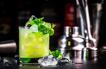 Green Negroni cocktail drink with gin, aperitif, liqueur, mint and ice. Black bar counter background with bottles