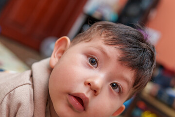A baby with messy hair and a blue eye. The baby is looking at the camera. The baby is wearing a brown sweater