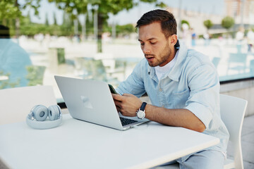 Young man focused on smartphone while working on laptop at outdoor cafe, illustrating multitasking and modern technology