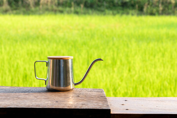Stainless steel gooseneck kettle on wooden table with green rice field background. Peaceful...