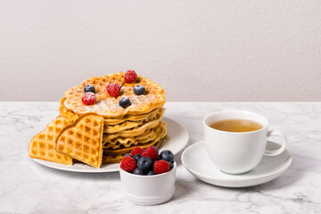 Stack of Belgian waffles, berries and cup of tea: delicious summer breakfast on marble table. Homemade waffle and mix of raspberries and blueberries as topping. Sweet dessert on white dishes