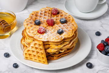 Stack of Belgian waffles with berries on top. Delicious summer breakfast from above. Heart-shaped waffle and mix of raspberries and blueberries as topping on marble background, top view