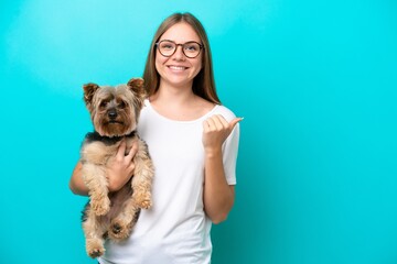 Young Lithuanian woman holding a dog isolated on blue background pointing to the side to present a product
