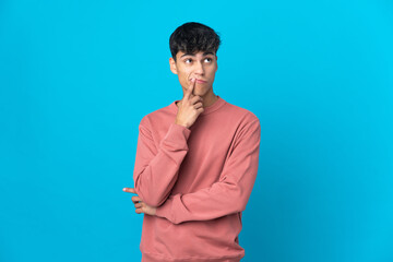 Young man over isolated blue background having doubts while looking up