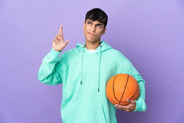 Young man playing basketball over isolated purple background with fingers crossing and wishing the best