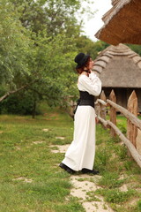 Young woman in old fashioned clothes standing near wooden fence at old country house