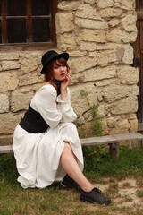 Old fashioned young woman sitting on a bench at ancient stone house