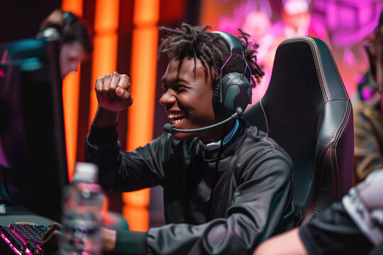 A triumphant African American pro gamer wearing a headset celebrates a win at an esports event, smiling and raising his fist in joy. Excited winner of cybersport competition