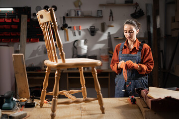 Young female carpenter wearing orange gloves working in workshop, restoring and renovating old wooden furniture