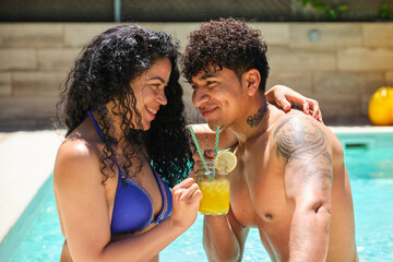 A couple looking into each other's eyes and smiling while holding a cocktail inside a pool. The man has a tattoo on his arm