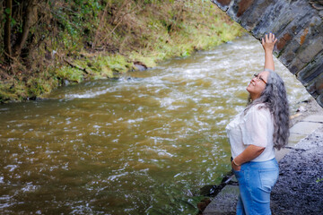 Senior adult woman standing in profile on bank of stream looking up, under stone bridge, Val-Dieu Abbey gardens, casual clothing, jeans and blouse, blurred background, cloudy day in Aubel , Belgium