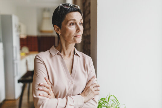 Portrait of worried anxious strict pretty mature woman with glasses on head standing at home looking through window with folded arms, being stressful because of future of her grandchildren