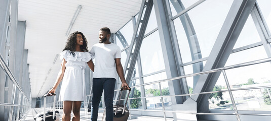 Cheerful black couple walks through an airport terminal together, both pulling their wheeled...