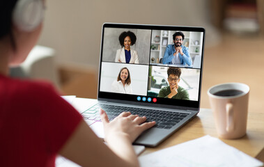 A woman sits at a desk, her back to the camera, with a laptop in front of her. She is participating in a video conference call with multiple participants who are visible on the laptop screen
