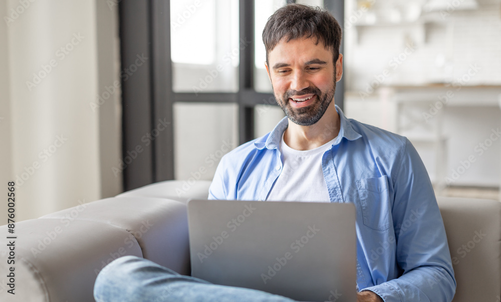 Poster a man is sitting on a couch at home, using his laptop. he is smiling and appears to be enjoying his 