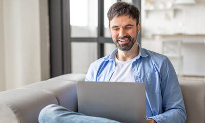 A man is sitting on a couch at home, using his laptop. He is smiling and appears to be enjoying his time online. The man is wearing a blue shirt and jeans, and the couch is white.