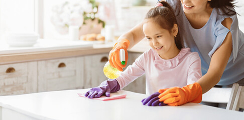 A young girl wearing pink shirt and purple gloves is cleaning a table in the kitchen while her...