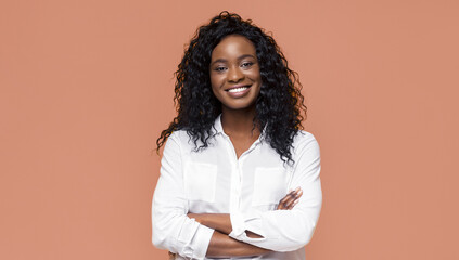 Confident Black woman with long, curly black hair. She is wearing a white button-up shirt and smiling directly at the camera with her arms crossed. She is standing in front of a peach background.