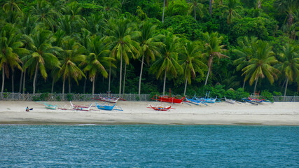 Shore of a tropical island. View from the sea of ​​boats on a sandy beach under palm trees.