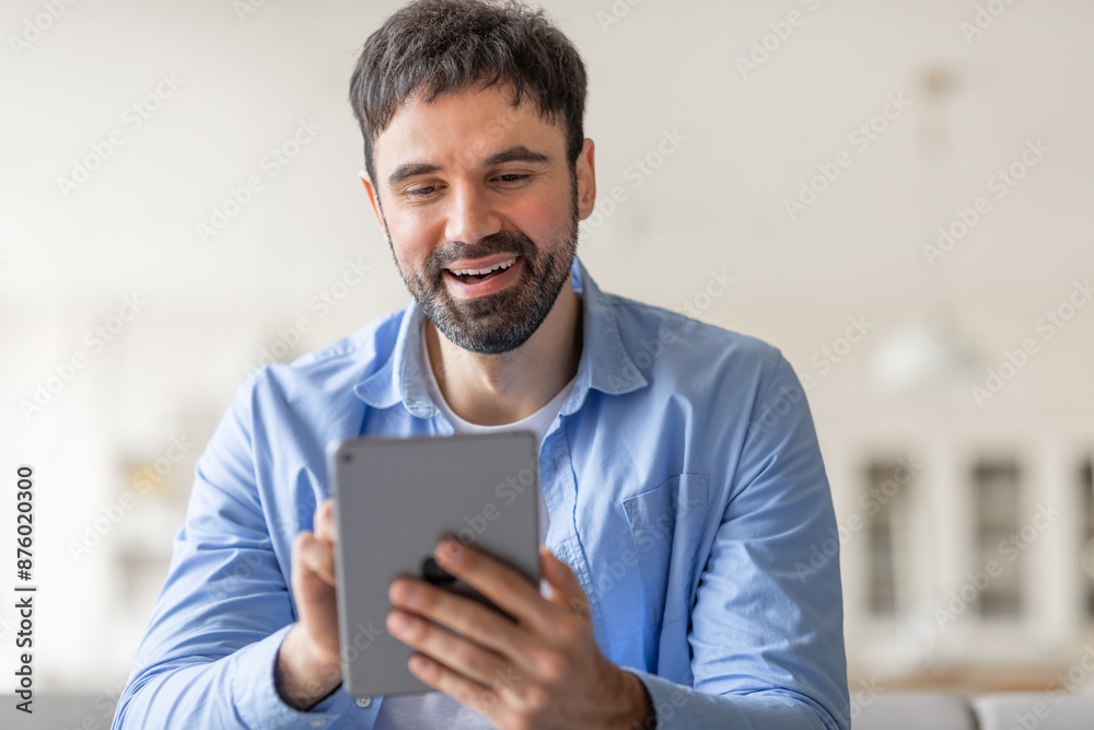 Poster A man is smiling as he uses a tablet. He is wearing a light blue button-down shirt and is sitting indoors. He appears to be enjoying his time using the device.