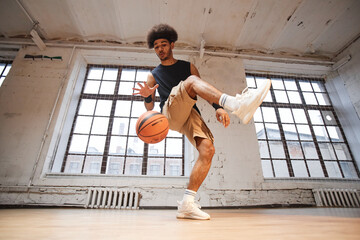 Wide angle action shot of young man as professional basketball player demonstrating tricks with ball while practicing in indoor gym copy space