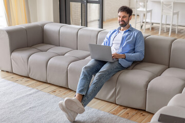 A man sits on a grey modular sofa in a modern living room, using a laptop computer. He is smiling and looks relaxed, possibly enjoying a break from work, high angle view