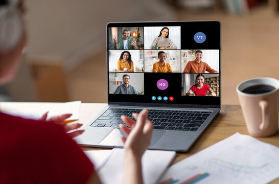 A woman sits at her desk, participating in a video conference call on her laptop. The screen displays multiple participants, with their names and initials visible