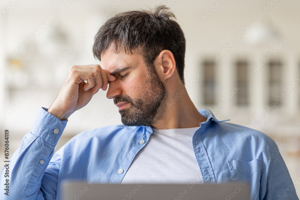 Poster tired man sitting in a chair inside, looking down and rubbing his eyes with his right hand. he appea
