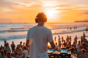 Fotobehang A DJ performs in front of a large crowd at a beach party during sunset. The sun is setting over the ocean, and the waves are crashing in the background. © MYDAYcontent