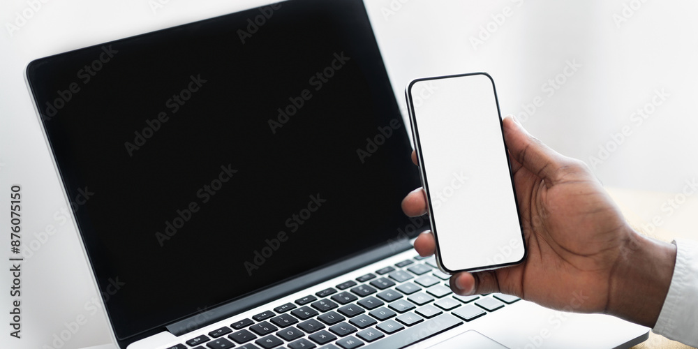 Poster a hand holds a smartphone with a blank screen near a laptop with a black screen.