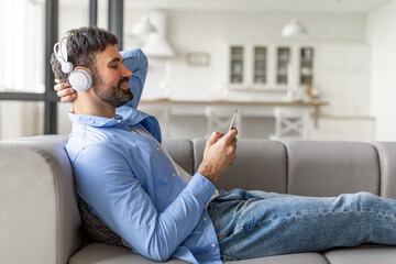 Fototapeta premium A man is sitting on a couch, wearing headphones and using his smartphone. He appears to be relaxed and enjoying his time. The man is wearing a light blue shirt and blue jeans