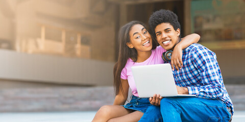 A teen black couple sits together on a ledge in the city, laughing and using a laptop. The girl is wearing a pink shirt and denim skirt, while guy wears a plaid shirt and jeans.