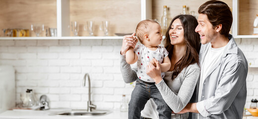 A young family of three stands in their modern kitchen, filled with white cabinets and brickwork. The mother holds their baby while the father looks on, beaming with joy, copy space - Powered by Adobe