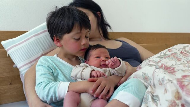 Older sibling holds the baby on their lap while mother watches, creating a heartwarming family moment. The older child&rsquo;s gentle touch and care for the baby reflect the close bond and affection