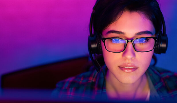 A woman wearing glasses and headphones is looking intently at a computer screen. The room is lit in a purple and blue color scheme, creating a vibrant and modern atmosphere