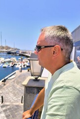 Young adult man, seen in profile looking at boats in the port, on a sunny day. Tourist in profile, looking at boats in the port of Las Galletas in Tenerife. Boy relaxing at the port looking at boats. 