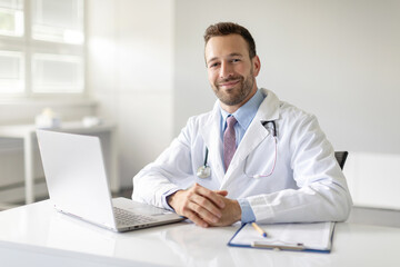 Portrait of professional man general practitioner working on laptop computer in clinic office, preparing to attend appointment, smiling at camera