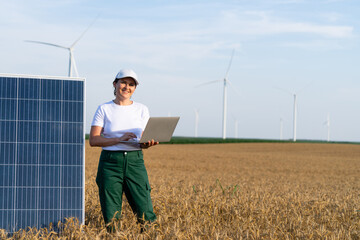 Woman farmer wearing white cap and t-shirt with laptop stands next to solar panel. Wind turbines in...