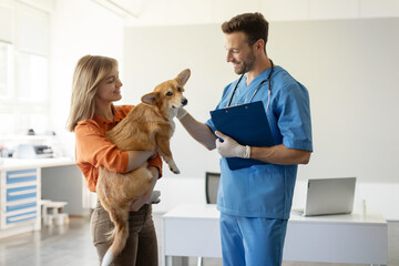 Happy woman owner holding her Pembroke Welsh Corgi dog during vet consultation, male veterinarian...