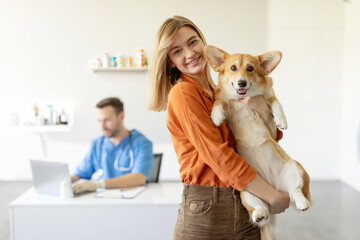 Happy woman holding her Pembroke Welsh Corgi dog, receiving care from male veterinarian in clinic, looking and smiling at camera