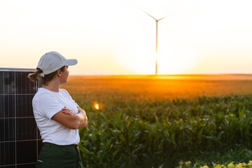 Woman wearing white cap stands next to solar panel at sunset. Wind turbines in the background