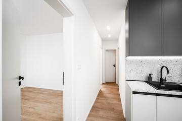 A modern kitchen with black upper cabinets, a granite countertop and backsplash, a black sink, wooden parquet flooring, and a view into an empty corridor and adjacent rooms.
